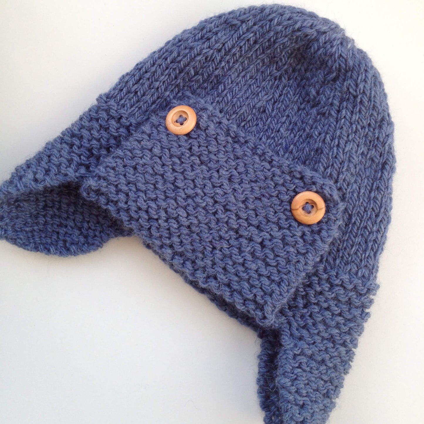 an aviator hat knitting pattern in child to adult sizes, shown laid flat and knitted in blue yarn