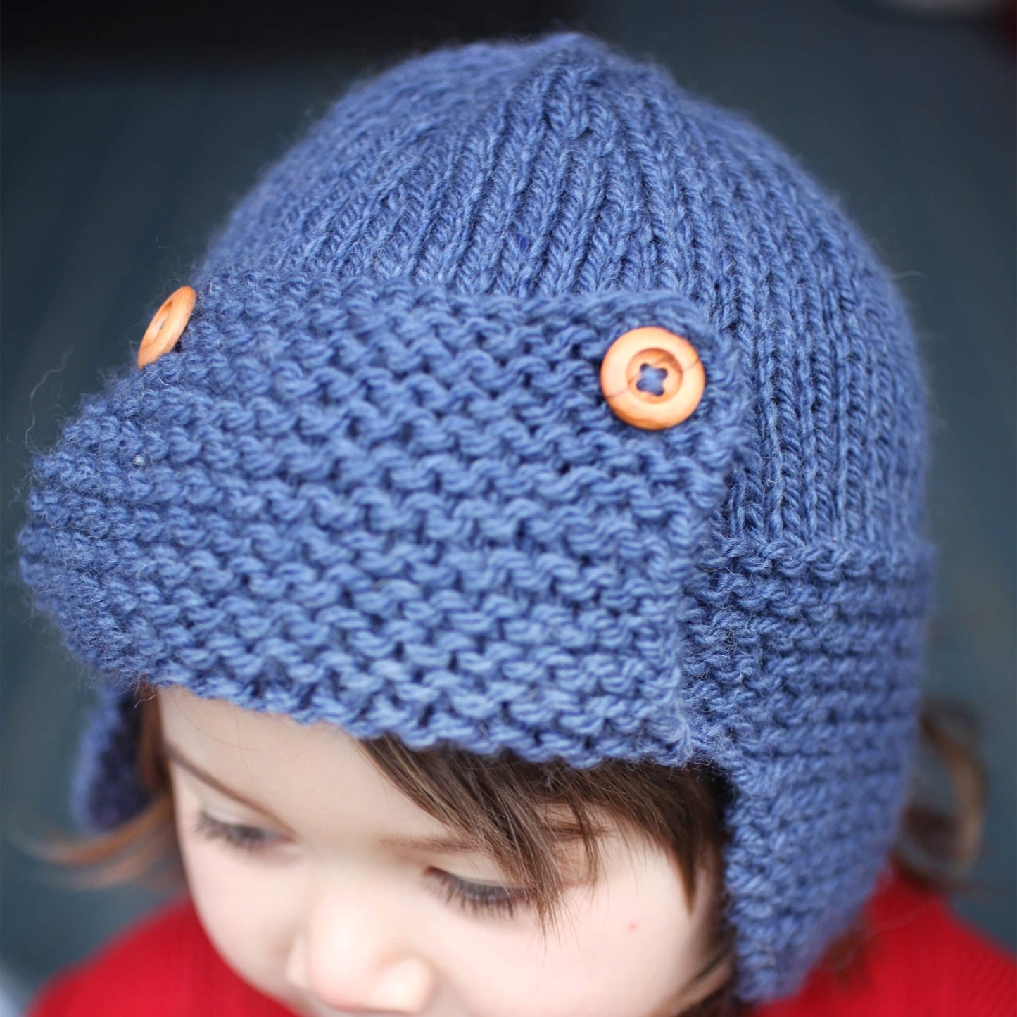 an aviator hat knitting pattern which uses chunky weight yarn shown in blue modelled on a little girl 