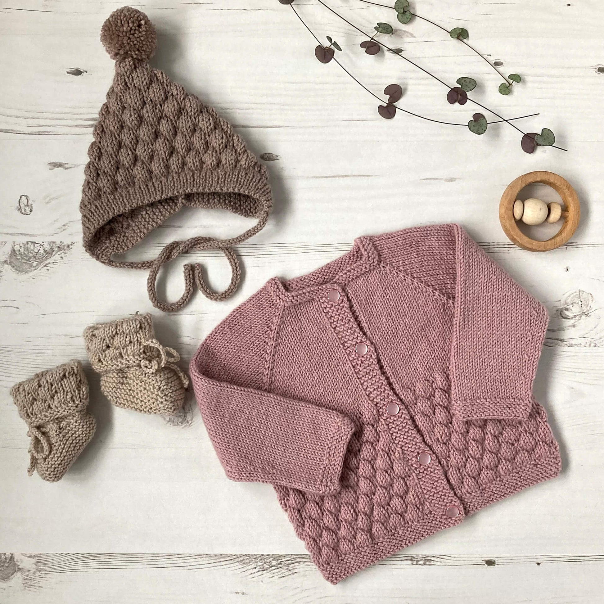 a baby bubble stitch knitting pattern. the pattern includes a cardigan shown in dusky pink , and booties and a pixie hat in shades of brown