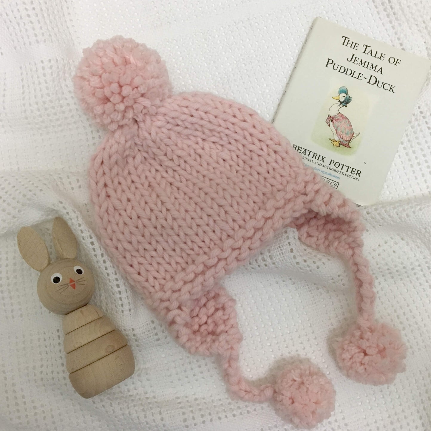 a baby earflap hat knitting pattern shown in pink on a blanket with baby toys