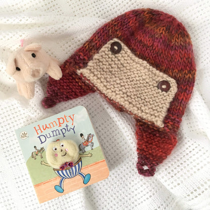 a flatlay of a baby knitting pattern for an aviator hat with button fastening styled with baby toys on a white blanket