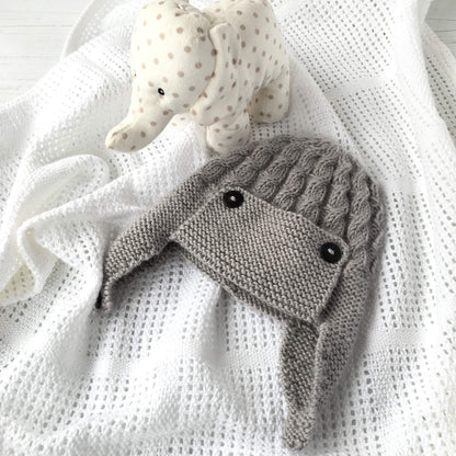 a baby aviator hat laying on a white blanket with toy nearby. the baby hat knitting pattern features a cable design and button fastening earflaps