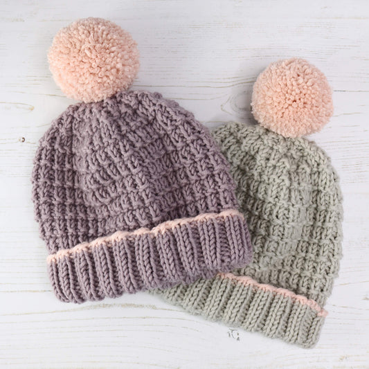 a knit hat pattern with sizes from baby to adult, samples shown are knit in lilac and grey, both with pink pompoms