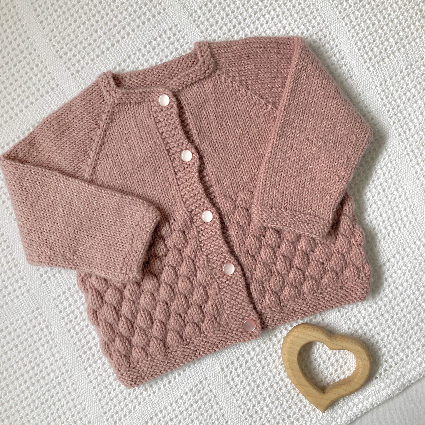 a bubble stitch cardigan knitting pattern for a baby shown in pink laying on a cotton blanket