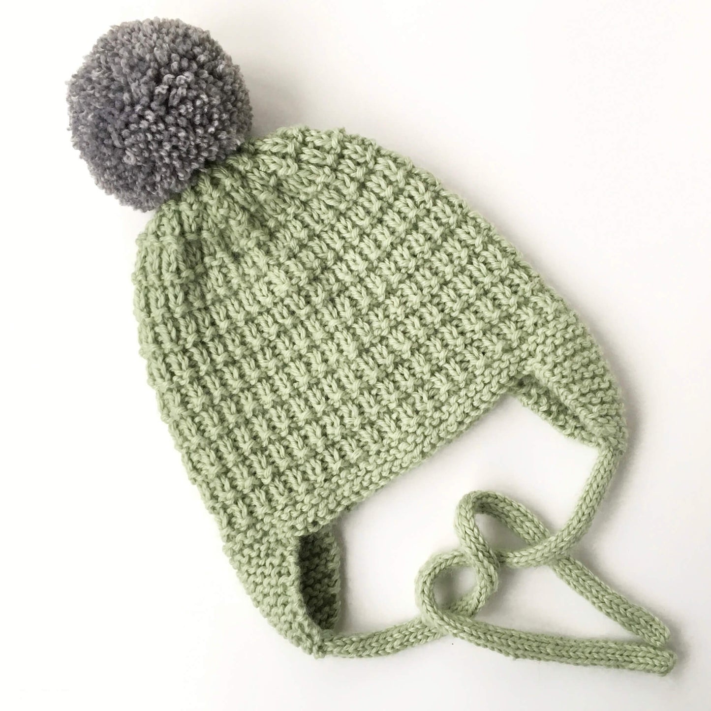 an earflap hat knit pattern for kids. shown in green yarn with a grey pompom