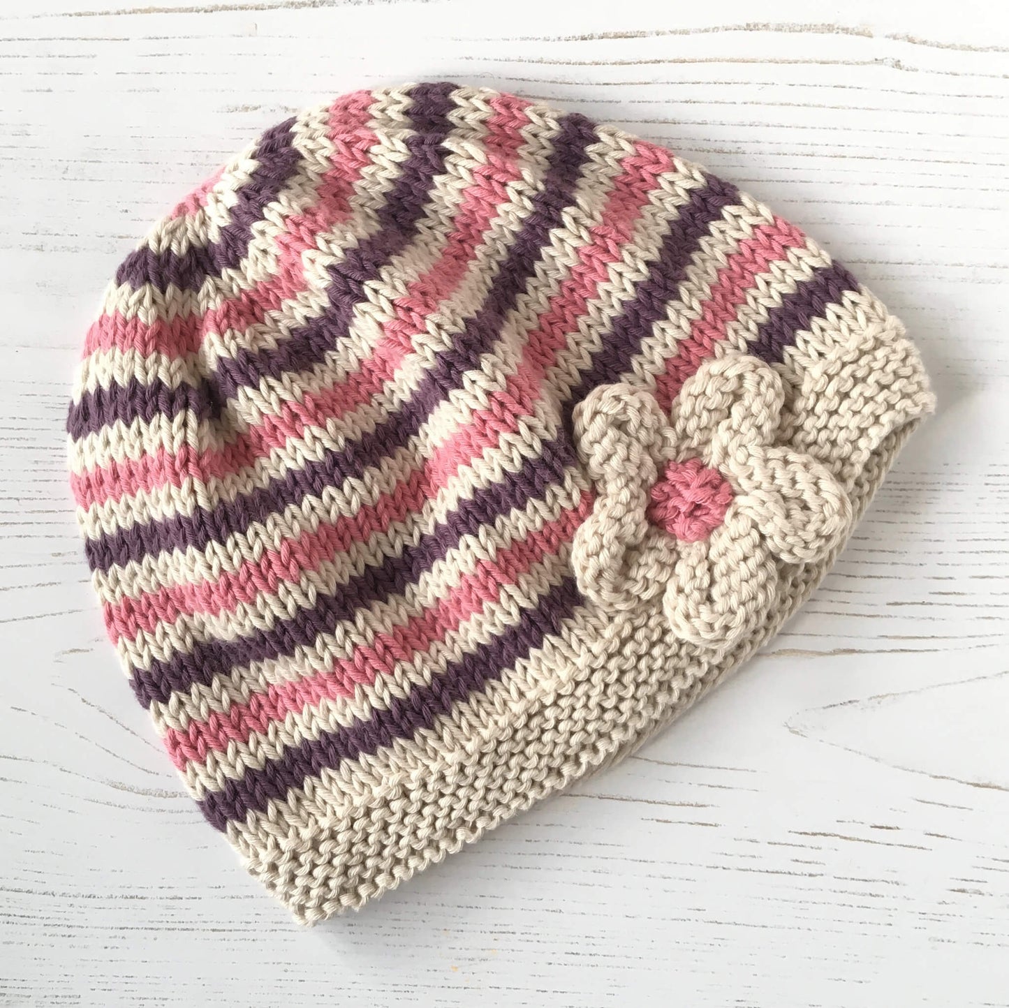 an easy baby hat knitting pattern. the hat has maroon and pink narrow stripes and a flower embellishment