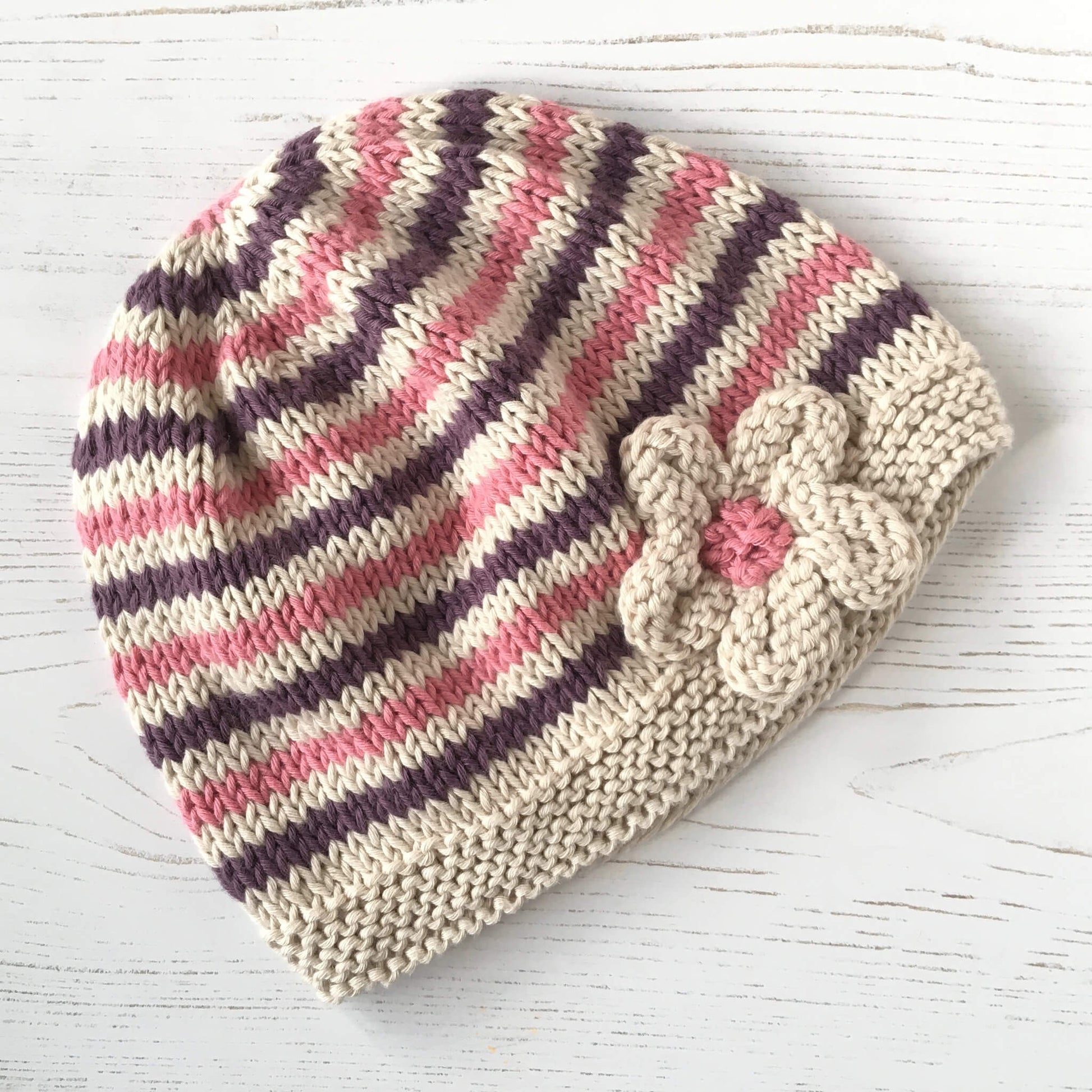 an easy baby hat knitting pattern. the hat has maroon and pink narrow stripes and a flower embellishment