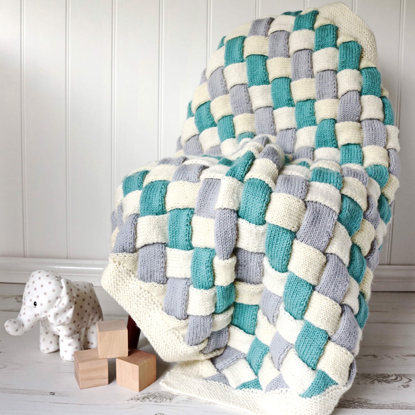 entrelac knitting pattern for a baby blanket draped over a chair with toys nnearby