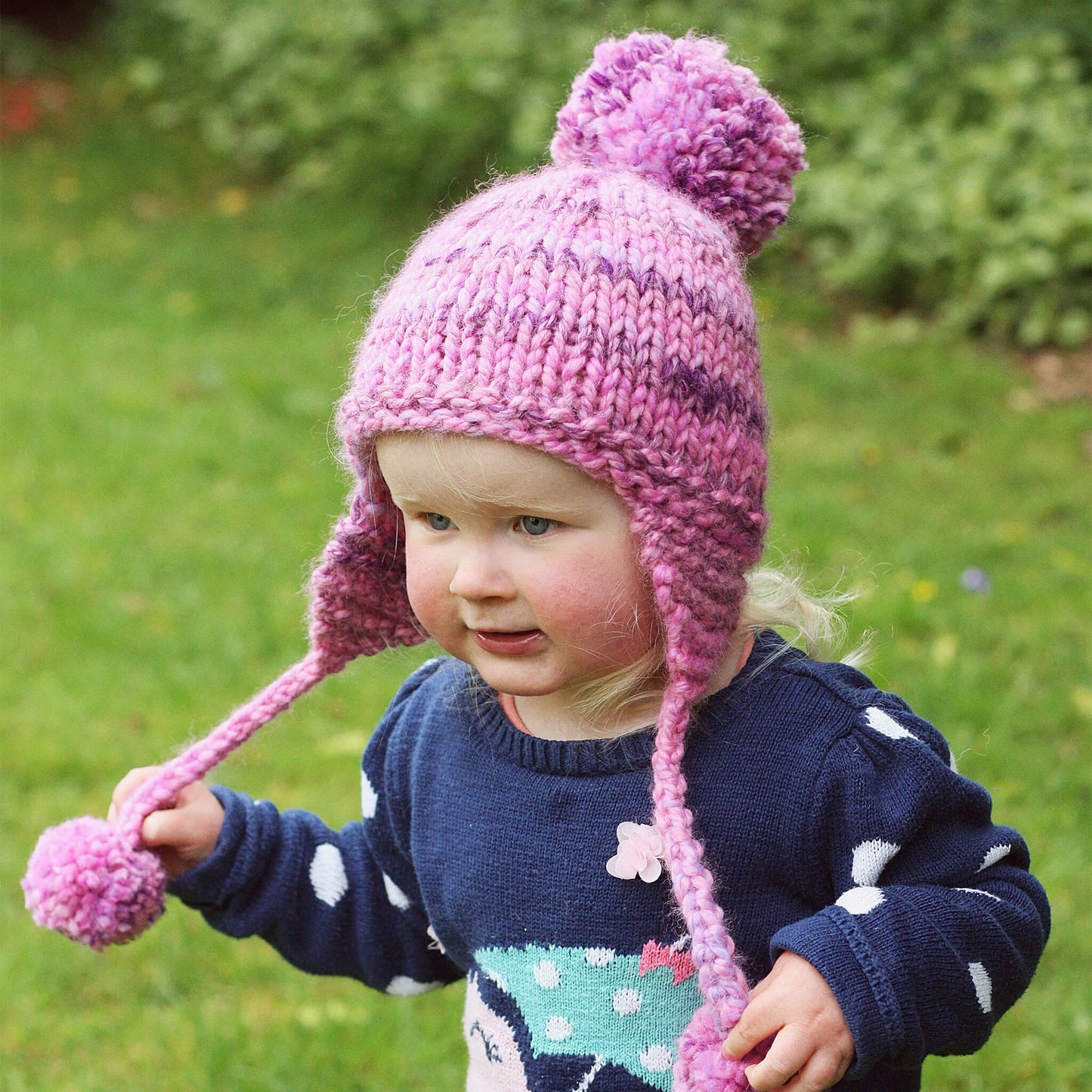 a kids hat knitting pattern modelled by a toddler running holding the pompoms