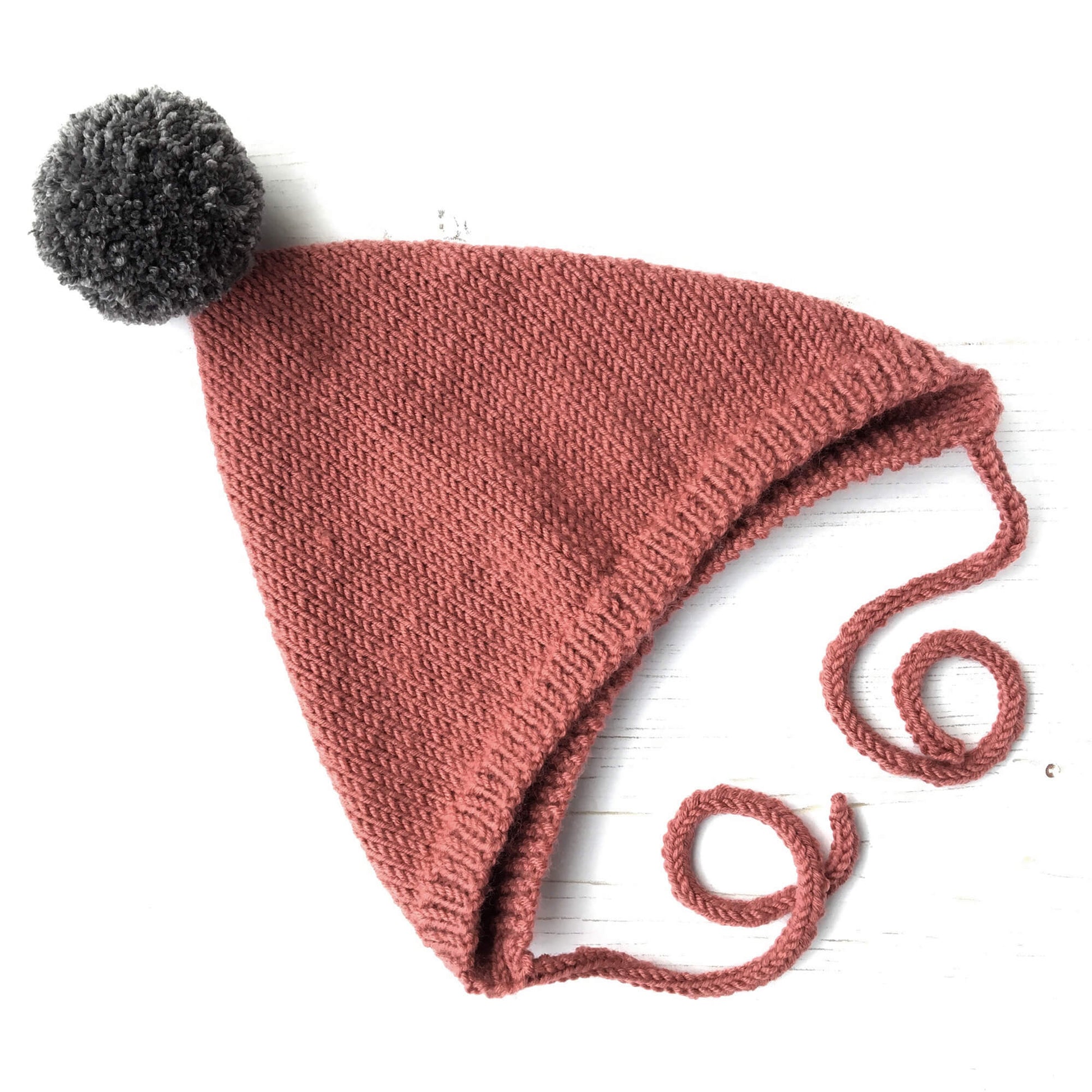 a knit hat pattern for a pixie hat. the hat is shown flat in rust colour yarn with a dark grey pompom