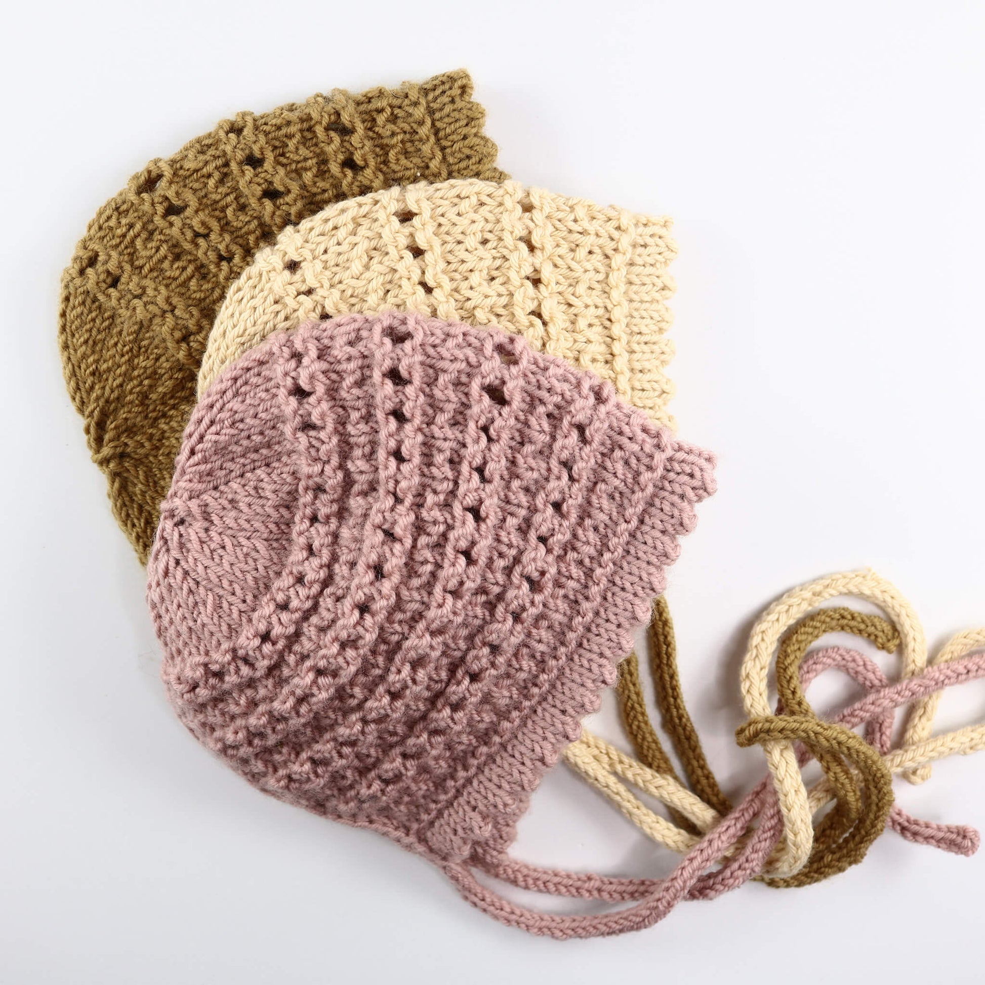 a knitting pattern for a baby bonnet. three samples are shown knit in pink, cream and mustard yarns