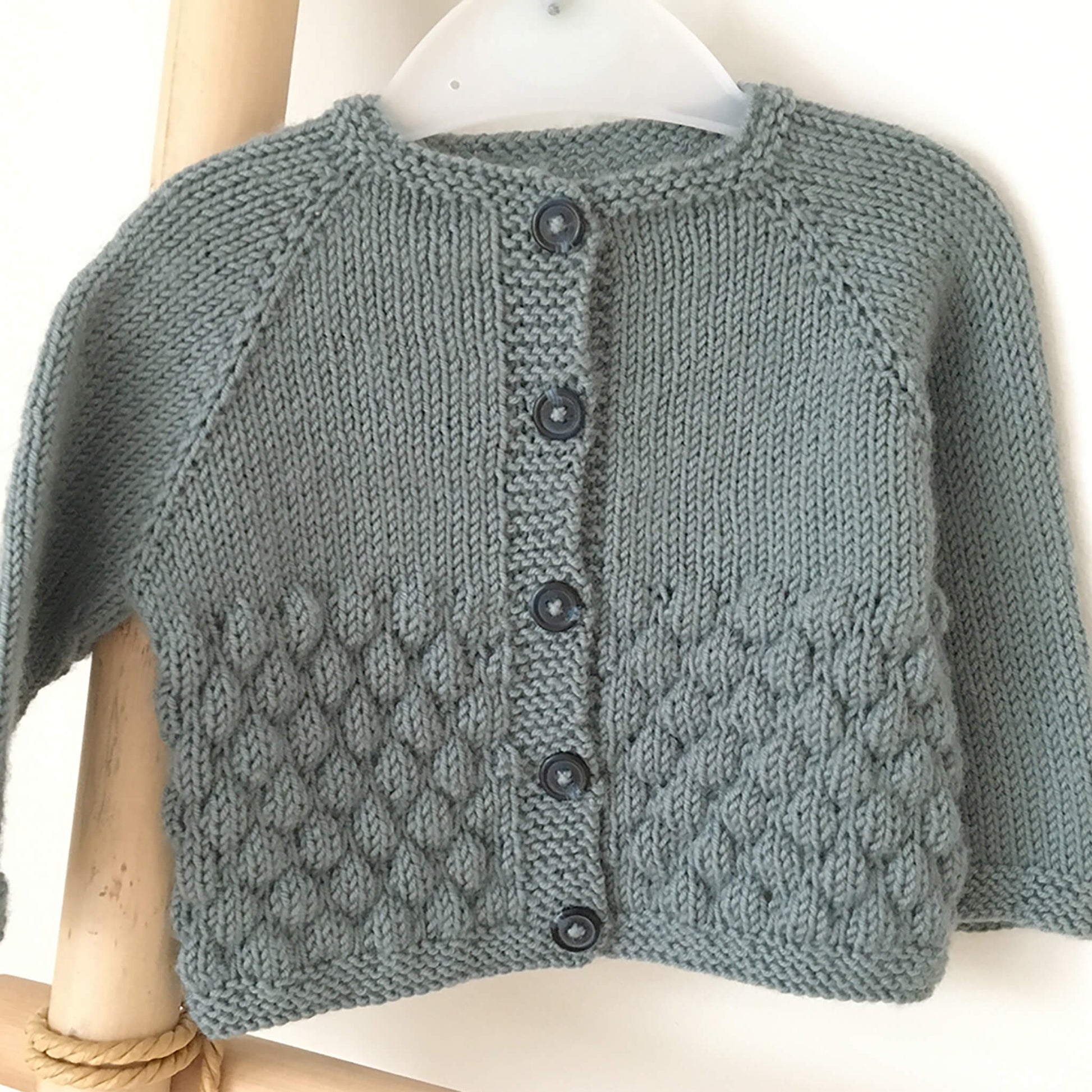 a knitting pattern for a baby cardigan. the grey-blue sample is photographed on a hanger