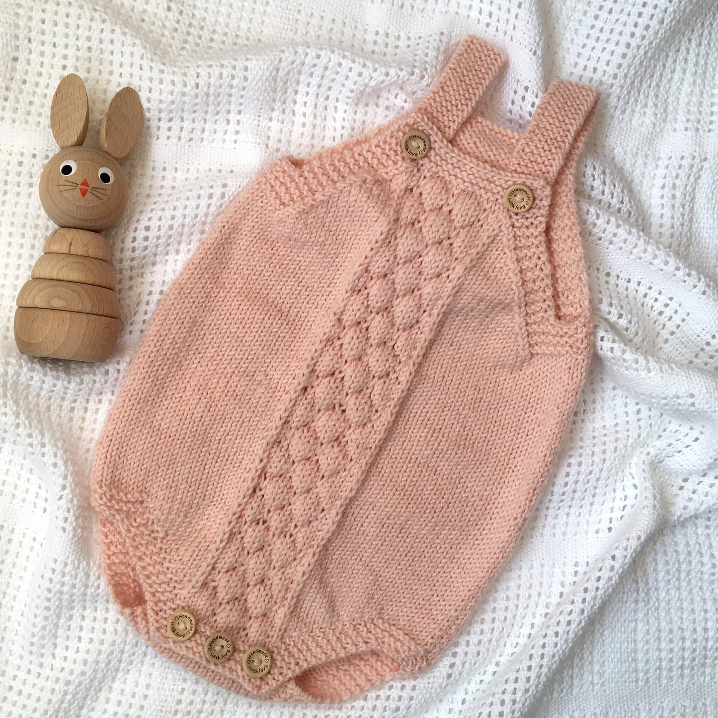 knitting pattern for a baby romper shown in pink laying on a white cotton blanket