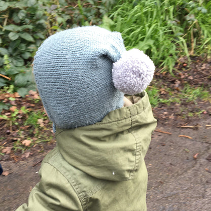 a knitting pattern for a pixie hat. photo shows the side view of a toddler wearing a light blue hat with grey pompom