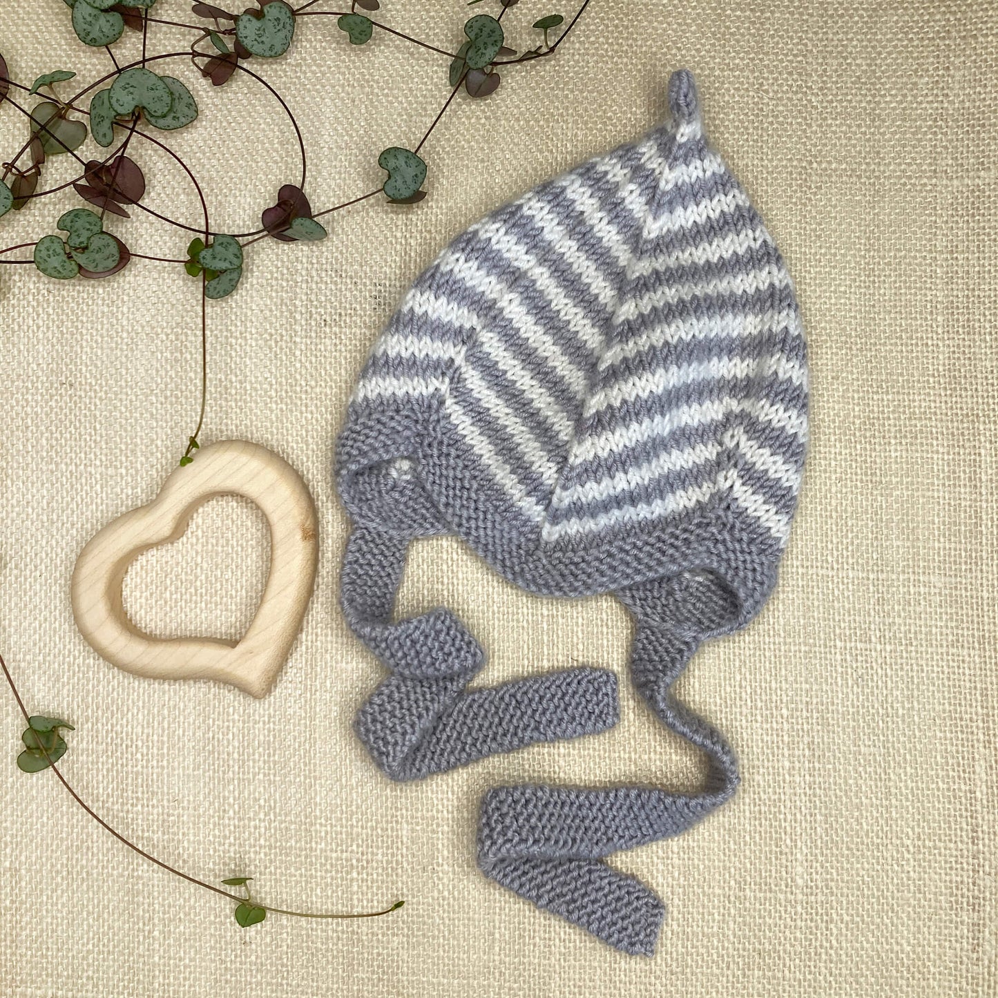 a flat lay showing a grey striped baby pixie hat knitting pattern on a hessian background with leaves and baby toy