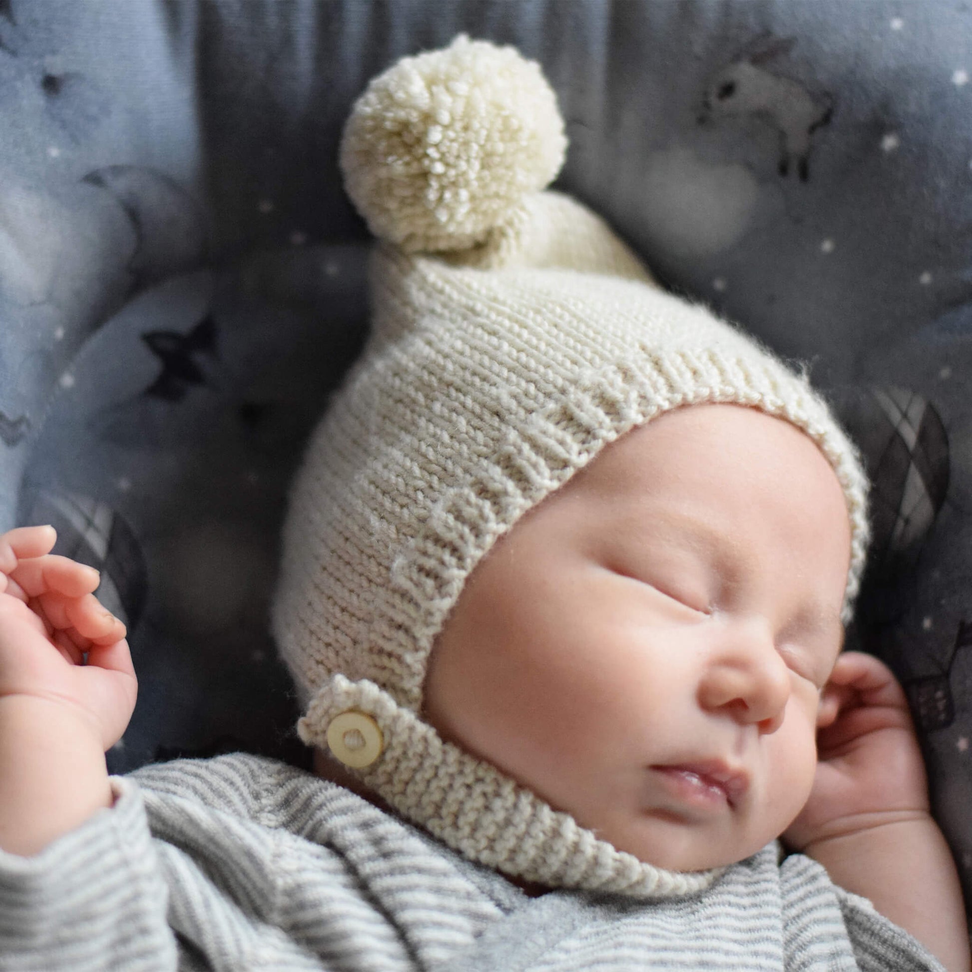 a simple knit pattern for a pixie hat. photo shows a sleeping baby wearing a cream colour hat with button fastening strap