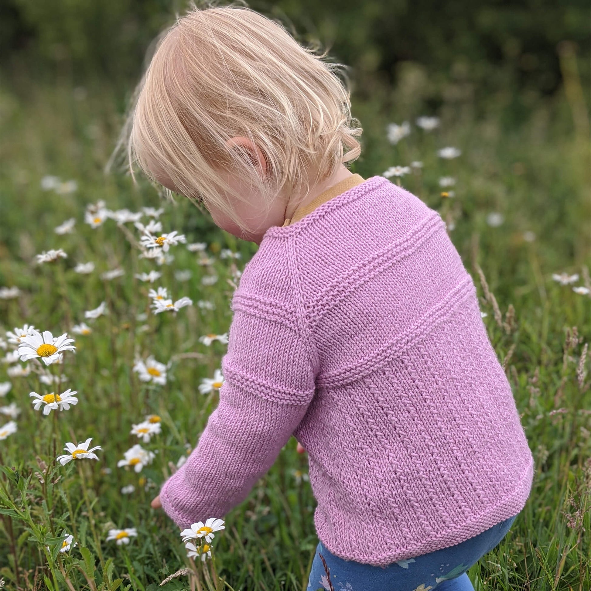 a cardigan knitting pattern that is worked from the top down. cardigan is modelled by a toddler crouching to pick flowers