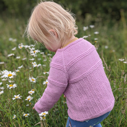 a cardigan knitting pattern that is worked from the top down. cardigan is modelled by a toddler crouching to pick flowers