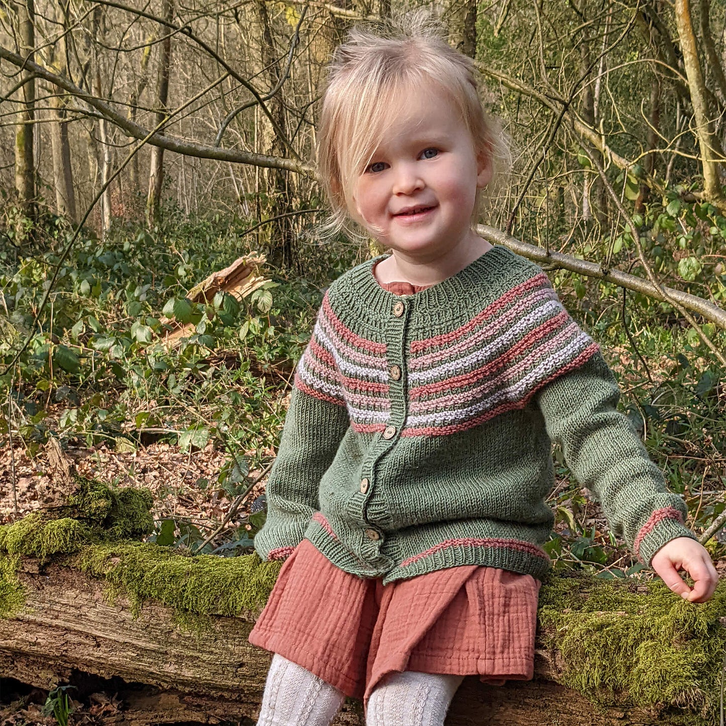 little girl sitting in woodland modelling a top down cardigan knitting pattern